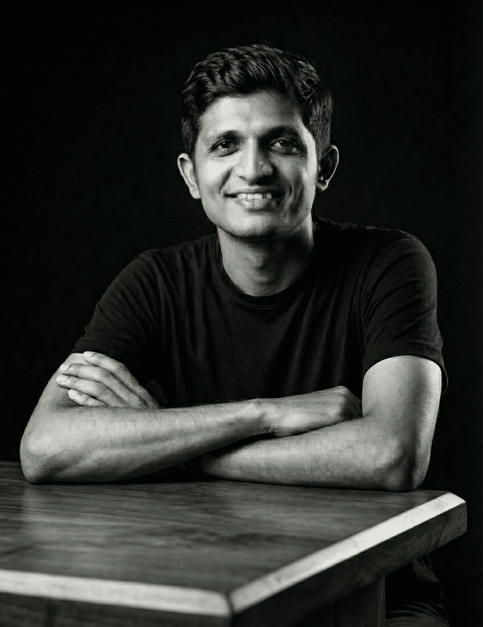 Black and white portrait of Shanwill Pinto smiling, arms folded on a wooden table